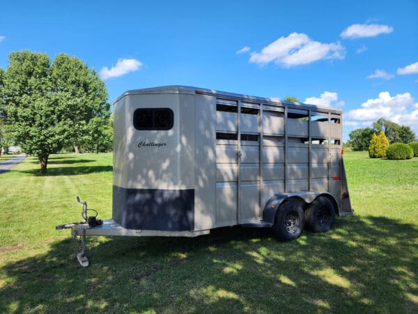 A horse trailer parked in the grass near some trees.